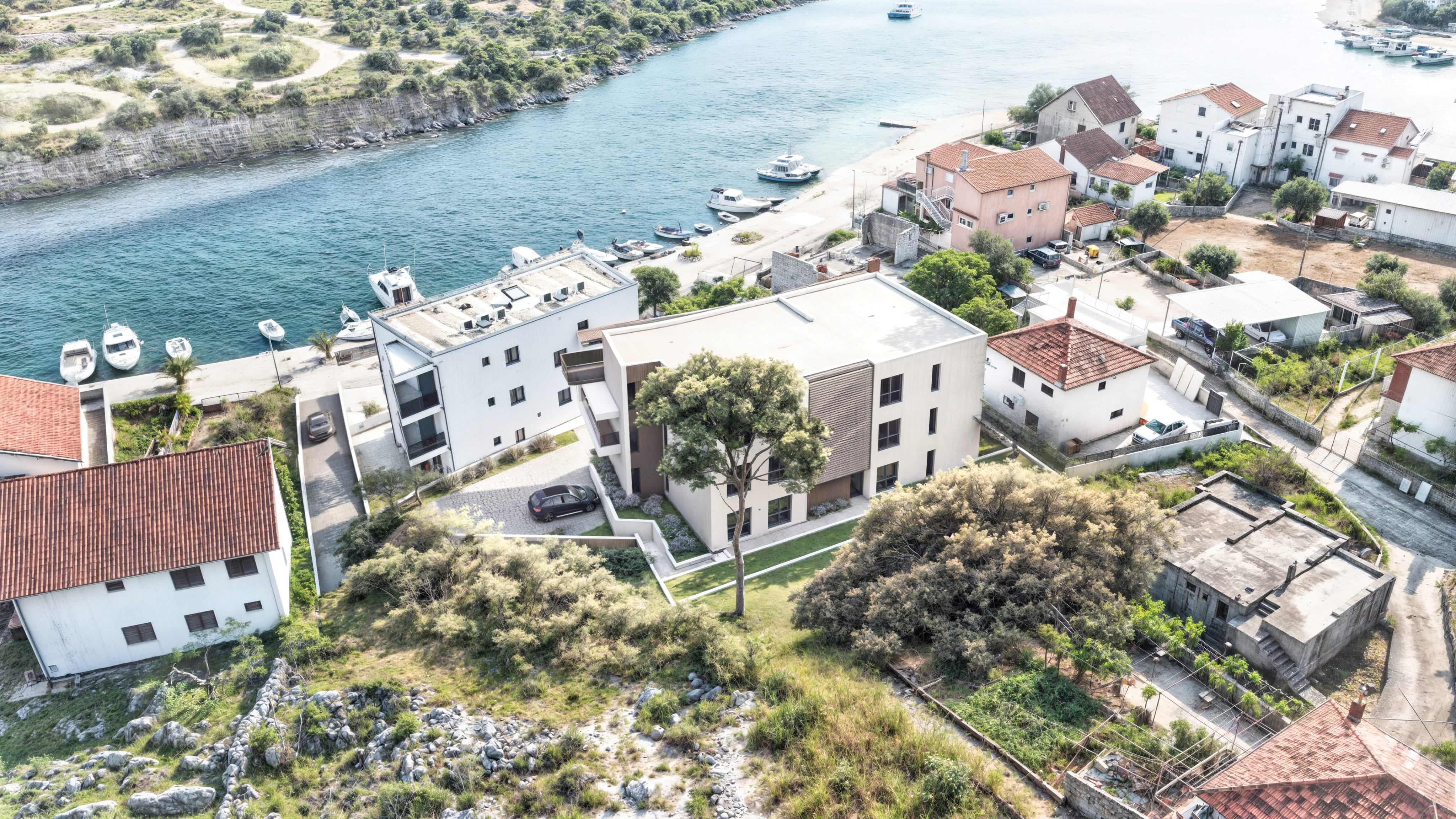 Zračna fotografija novogradnje u Rogoznici, smještene uz more, s pogledom na uvalu i blizinom plaže i rive s brodicama.